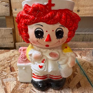 May include: A ceramic cookie jar shaped like a nurse doll with bright red yarn-style hair, a white nurse cap with a red cross, a white uniform with a red heart on the pocket, red and white striped socks, and black shoes, holding a spoon.