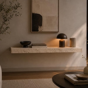 Può includere: Una lunga mensola rettangolare sospesa in pietra beige chiaro dalla superficie grezza. La mensola è fissata alla parete e ospita una ciotola marrone scuro, due libri sovrapposti, una piccola lampada marrone scuro a forma di fungo e un vaso cilindrico scanalato.