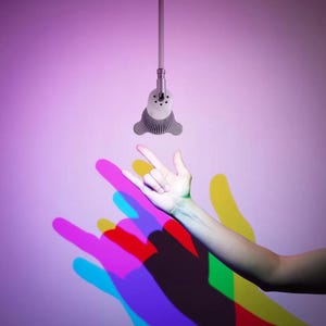 May include: A hand forming the 'rock on' gesture, casting colourful shadows on a purple wall. Above, a white and grey light fixture is suspended from the ceiling.