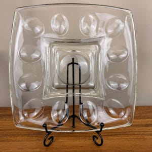 May include: A square, clear glass serving platter featuring a central circular indentation surrounded by eight smaller, evenly spaced circular indentations along the perimeter. The platter is supported by a black metal wire display stand.