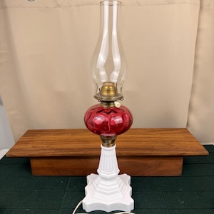 Puede incluir: Una lámpara de mesa con base de vidrio lechoso blanco sobre un pedestal cuadrado escalonado. La sección central es un depósito redondo de vidrio rojo con textura de hoyuelos. Incluye un quemador de latón y una pantalla de chimenea de vidrio transparente.
