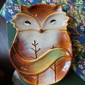 Op de afbeelding: Een keramieken schaal in de vorm van een vos met een glanzende afwerking. De vos heeft een bruin en crème kleurenpalet met een opvallende staart die over het onderste gedeelte loopt. Twee kleine takvormige details zijn zichtbaar.