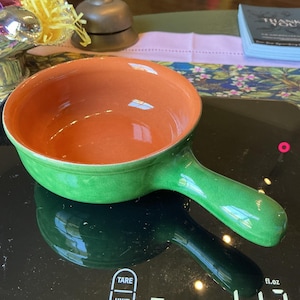 May include: A round ceramic bowl with a single handle, featuring a glossy green exterior and a smooth orange interior. The surface of the green glaze shows a fine crackle pattern. The bowl is positioned on a black digital scale with visible text.
