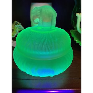 Op de afbeelding: Een gloeiende neon-groene glazen pot met een deksel voorzien van een gebeeldhouwd olifantje bovenop. De pot heeft een afgeronde, geribbelde basis en een getextureerd dekseloppervlak dat een helder fluorescerend licht uitstraalt.