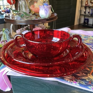 Könnte beinhalten: Eine leuchtend rote Glassuppenschale mit zwei seitlichen Griffen, die auf einer passenden roten Glasuntertasse steht. Das Set zeichnet sich durch eine glatte, durchscheinende Oberfläche und ein kreisförmiges Design in tiefem Karmesinrot aus.