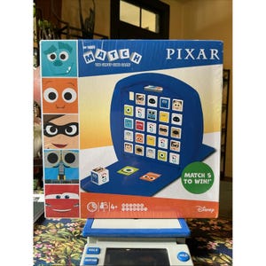 Può includere: Una scatola del gioco da tavolo Top Trumps Match Pixar con un tabellone a griglia blu e vari cubi personaggio. La confezione mostra il testo Match The Crazy Cube Game, Pixar e Match 5 To Win. La scatola include illustrazioni colorate di personaggi.