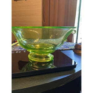 May include: A translucent lime green glass bowl with a wide, flared rim and a pedestal base. The bowl features small, decorative glass loops on opposite sides of the rim. The glass has a smooth, polished finish and a vibrant, light-catching color.