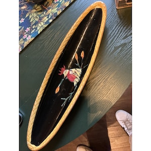 May include: An elongated, oval-shaped ceramic tray with a glossy black interior and a textured, light tan braided border. The center of the tray features a painted illustration of a rooster with red, white, and yellow plumage among green stems.