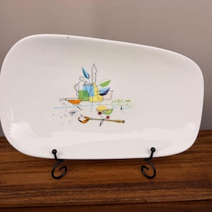 Op de afbeelding: Een gebroken witte rechthoekige serveerschaal met afgeronde hoeken, voorzien van een centrale grafische illustratie van keukenartikelen, waaronder een karaf, een kom, een lepel en een schijfje citroen in blauwe, groene, gele en zwarte lijntekeningen.