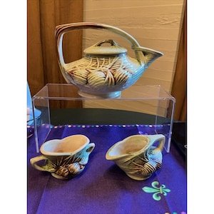 Può includere: Una teiera in ceramica con coperchio e due piccole brocche abbinate, tutte caratterizzate da una smaltatura verde chiaro con accenti marroni e un motivo in rilievo di pigne e aghi. La teiera ha un manico alto e un beccuccio.