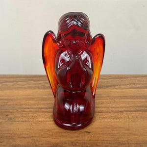 May include: A translucent ruby red glass figurine of a kneeling angel with hands pressed together in prayer. The figure features molded hair, facial details, and large wings that transition into a lighter orange hue at the outer edges.