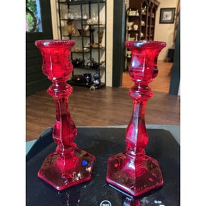 May include: A pair of matching ruby red glass candle holders featuring a faceted hexagonal base and a slender, tapered stem that widens into a flared top. The translucent red glass reflects light, showcasing a smooth, polished surface finish.