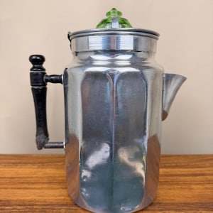 May include: A metallic silver percolator coffee pot with a faceted, multi-sided body. It features a black handle, a short spout, and a lid topped with a translucent, light green glass knob. The surface shows subtle signs of wear and use.