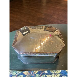 May include: A polished silver-toned metal box with an octagonal shape. The lid features a smooth, reflective surface with a decorative beaded edge detail along the perimeter. The sides of the box are straight and maintain the octagonal form.