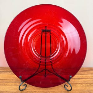 Puede incluir: Un plato circular de vidrio rojo translúcido con una superficie lisa y pulida, un borde ancho y plano y un centro hundido. El plato se exhibe en posición vertical sobre un soporte de alambre de metal negro, mostrando su color vibrante.