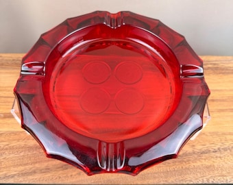 Könnte beinhalten: Ein runder Aschenbecher aus durchscheinendem rotem Glas mit einem gewellten, facettierten Rand und vier deutlichen kreisförmigen Vertiefungen auf dem Innenboden. Das Design weist vier gleichmäßig verteilte Einkerbungen am Außenrand auf.