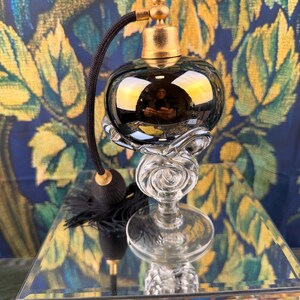 May include: A vintage-style perfume atomizer featuring a dark, reflective glass bulb with a gold-toned metal cap. The base is made of clear, sculpted glass with a swirling design. A black fabric-covered hose connects to a black tassel pump.