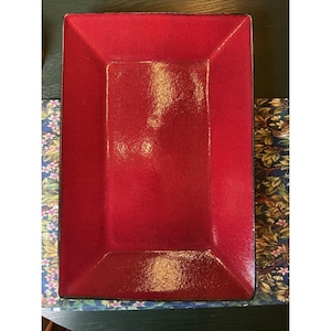 May include: A rectangular serving tray with a vibrant, glossy red finish and a dark, contrasting rim. The surface features a smooth, reflective texture with subtle variations in the red hue, creating a deep, rich appearance across the flat base.