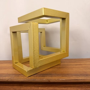 May include: A gold-colored metal sculpture featuring an impossible cube design with interconnected square frames. The object has a smooth, matte finish and consists of rigid, straight bars joined at right angles to create a geometric shape.