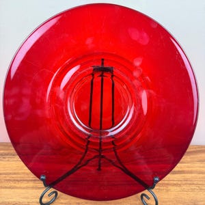 Puede incluir: Un plato circular de vidrio rojo translúcido con una superficie lisa y pulida, un borde ancho y un centro ligeramente hundido. El plato se exhibe en posición vertical sobre un soporte de alambre de metal negro, mostrando su color rojo intenso.
