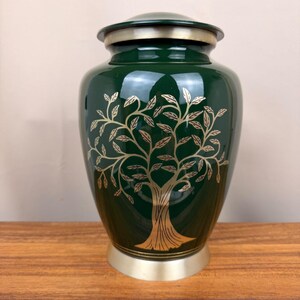 May include: A dark green metal cremation urn featuring a gold-toned tree of life engraving on the front. The urn has a smooth, glossy finish, a rounded gold-toned lid, and a matching gold-toned base, creating a balanced and cylindrical shape.