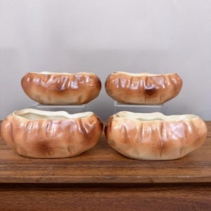 May include: Four ceramic bowls shaped like baked potatoes, each featuring a textured, light brown exterior with darker brown shading to mimic a crust. The bowls have an open, irregular top edge and a smooth, cream-colored interior surface.