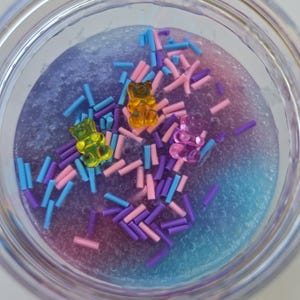 May include: A clear jar filled with colorful slime. The slime is a mix of blue, purple, and pink hues, speckled with small, colorful sprinkles. Three gummy bear candies in yellow, green, and purple are scattered throughout the slime.