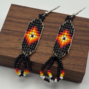 May include: A pair of dangle earrings featuring a rectangular beaded pattern in black, red, orange, yellow, and white. The earrings have silver-toned metal hooks and a fringe of black, red, orange, yellow, and white beads at the bottom.