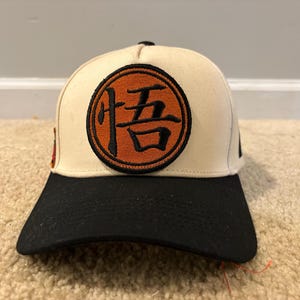 May include: A baseball cap with a cream-colored crown and a black brim. The front features a circular orange patch with a black embroidered Japanese kanji character. A small red and black rectangular emblem is visible on the side of the cap.