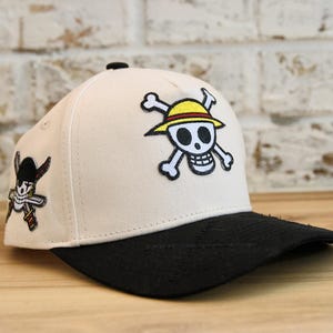 Pode incluir: Boné de beisebol branco com aba preta. O boné apresenta um logotipo de caveira e ossos cruzados com um chapéu amarelo e um personagem de pirata de desenho animado. O boné é feito de tecido.