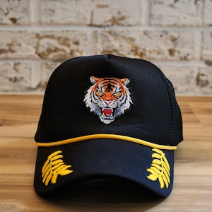 Puede incluir: Gorra de camionero negra con un rostro de tigre bordado en la parte delantera. La gorra tiene un ribete amarillo y diseños de hojas amarillas en la visera. La parte trasera es de malla.