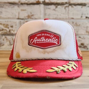 May include: Red and white trucker hat with the words "100% Original & Authentic" in a red and white emblem. The hat has gold leaf accents on the brim.
