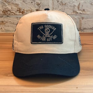 May include: A beige baseball cap with a black brim. The front features a rectangular patch with the words "The Goonies Never Say Die" and a skull and crossbones design. The cap is sitting on a wooden surface.