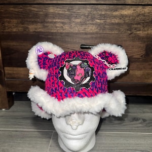 Puede incluir: Un gorro de ganchillo con un patrón rosa y morado vibrante, ribete de piel sintética blanca y dos orejas redondeadas. Un parche central muestra una cabeza de gato estilizada dentro de un engranaje negro. Incluye una cadena blanca y la palabra CRANKOAT.