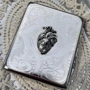 May include: A rectangular silver-toned metal case featuring an intricate engraved scrollwork pattern across the surface. Centered on the front is a three-dimensional, dark silver-toned anatomical heart emblem with detailed textures and veins.