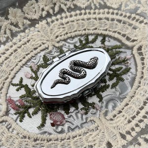 May include: A small, oval-shaped silver-toned metal pill box featuring a raised, textured snake emblem on the lid. The container has a hinged closure and a polished, reflective surface, showcasing intricate detailing on the serpent's body.