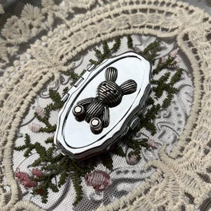 May include: A small, oval-shaped silver-toned metal compact mirror featuring a raised, textured rabbit figure on the lid. The compact has a polished, reflective surface and a hinged design, showcasing a detailed, ribbed pattern on the rabbit's body.