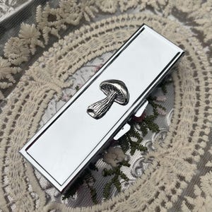 May include: A rectangular silver-toned metal case with a polished, reflective finish. A three-dimensional silver-toned mushroom emblem is attached to the centre of the lid. The case features a hinged opening mechanism on the side.