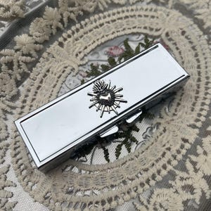 May include: A rectangular silver-toned metal lipstick case with a polished, reflective finish. The centre of the lid features a raised, three-dimensional silver-toned emblem of a sacred heart with radiating sunburst rays extending outwards.