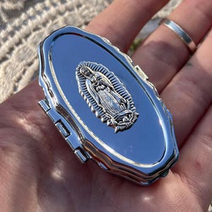 May include: A small, oval-shaped silver-toned metal pill box with a polished, reflective surface. The lid features a raised, detailed relief of the Virgin of Guadalupe centred on the top. The container has a hinged side closure mechanism.
