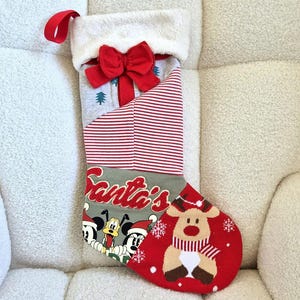 May include: A Christmas stocking featuring a white faux fur cuff with a red bow. The body has red and white stripes, a grey section with the text Santa's, and a red toe area with a reindeer graphic and Disney characters Pluto and Minnie.