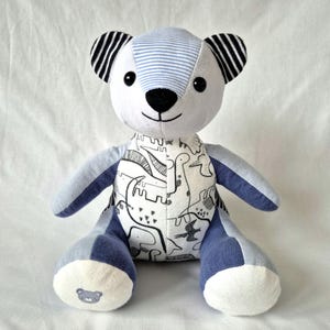 May include: A stuffed bear toy constructed from various fabric patches. The head features light blue and white stripes, while the ears are black and white striped. The torso displays a white fabric with black line drawings of dinosaurs.