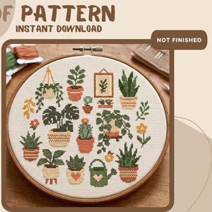 Op de afbeelding: Een kruissteekpatroon in een houten borduurring met diverse potplanten, hangend groen, een ingelijste botanische print en een kleine gieter. Het ontwerp gebruikt groen, oranje en geel garen op witte stof. Tekst luidt PDF PATTERN.