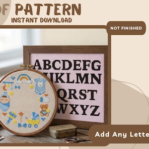 May include: A PDF pattern for cross-stitch featuring a circular hoop with baby-themed motifs including a swaddled infant, teddy bear, baby carriage, and rainbow. Beside it, a framed alphabet sampler with black letters on a light pink background.