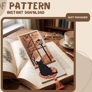 May include: A cross-stitch bookmark pattern featuring a black cat sitting on a round cushion, looking out a window at a street lamp and autumn trees. The rectangular design includes a brown tassel at the bottom. Text reads PDF PATTERN INSTANT DOWNLOAD NOT FINISHED.