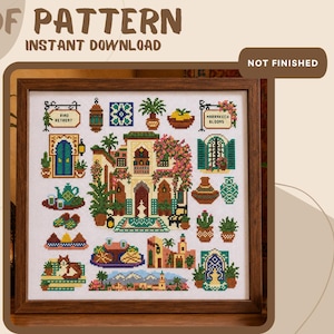 May include: A cross stitch pattern featuring a central building with pink flowers, surrounded by smaller motifs including a cat, a teapot, cacti, lanterns, and signs reading Riad Retreat and Marrakech Blooms, all framed in a dark wood frame.