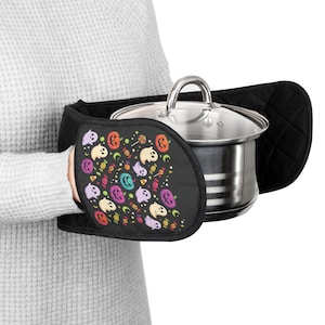 May include: A black double oven mitt featuring a colorful pattern of ghosts, jack-o'-lanterns, and candy. The mitt is quilted and holds a stainless steel pot with a glass lid. The design includes orange, purple, and yellow accents on a black background.