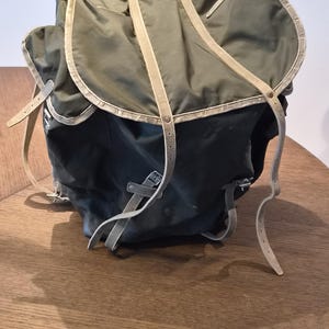 May include: An olive green backpack featuring a large top flap with light tan leather trim and matching adjustable leather straps. The bag includes a main compartment with a drawstring closure and side pockets, all secured with metal buckles.