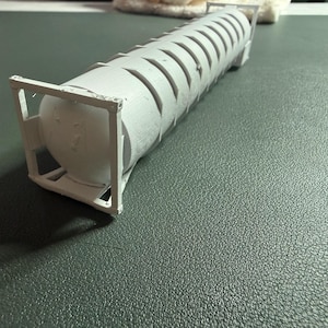 May include: A white 3D printed model of a cylindrical tank container featuring a ribbed exterior design. The tank is secured within a rectangular frame structure at both ends, showcasing a detailed industrial aesthetic with visible print layers.
