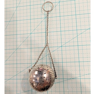 Puede incluir: Un infusor de té de metal plateado con un cuerpo esférico perforado diseñado para preparar té en hebras. El infusor está unido a una delicada cadena de metal que se conecta a un anillo circular en la parte superior, facilitando su manejo.